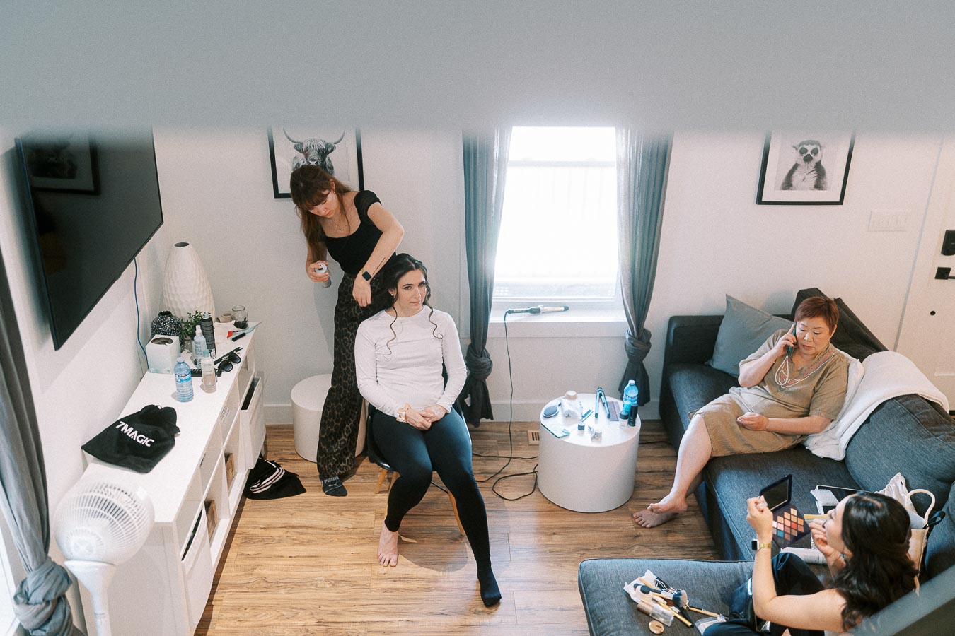 Bride sits in a bright room while a hairstylist curls her hair and others relax nearby during wedding preparations.