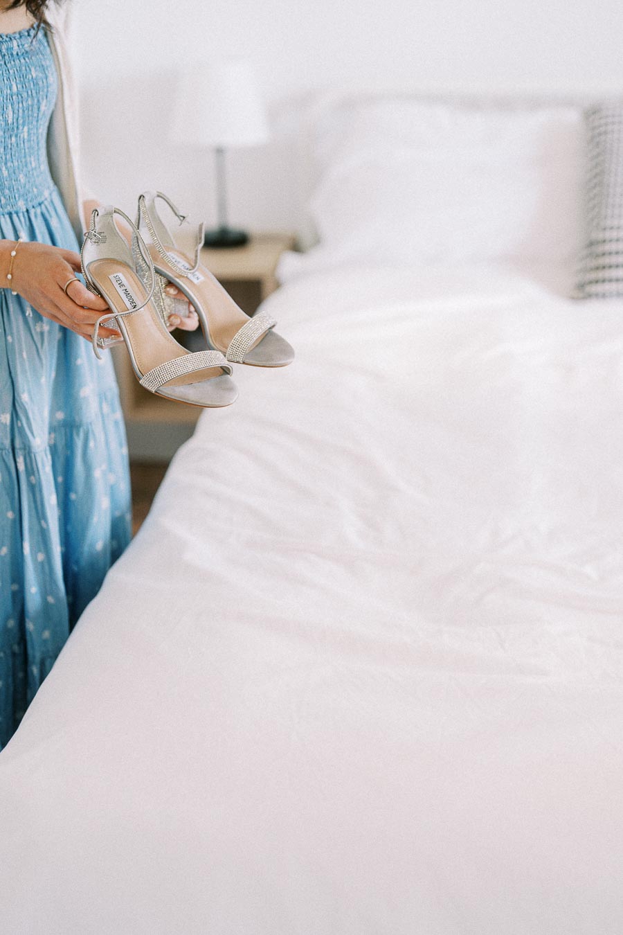 Hands holding silver Steve Madden bridal heels beside a white bed during wedding preparations