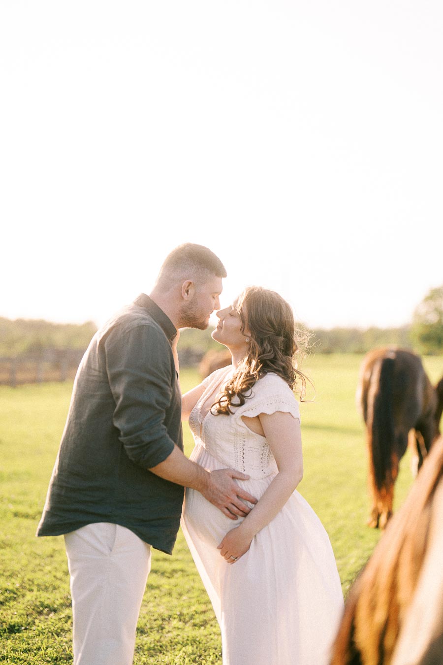 Expectant couple kissing in a field with horses nearby at The Green Acres