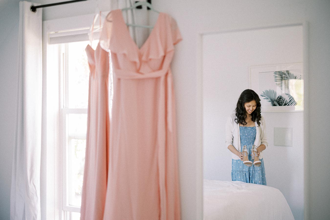 Woman holding sandals in a bright bedroom with a blush dress hanging in the foreground