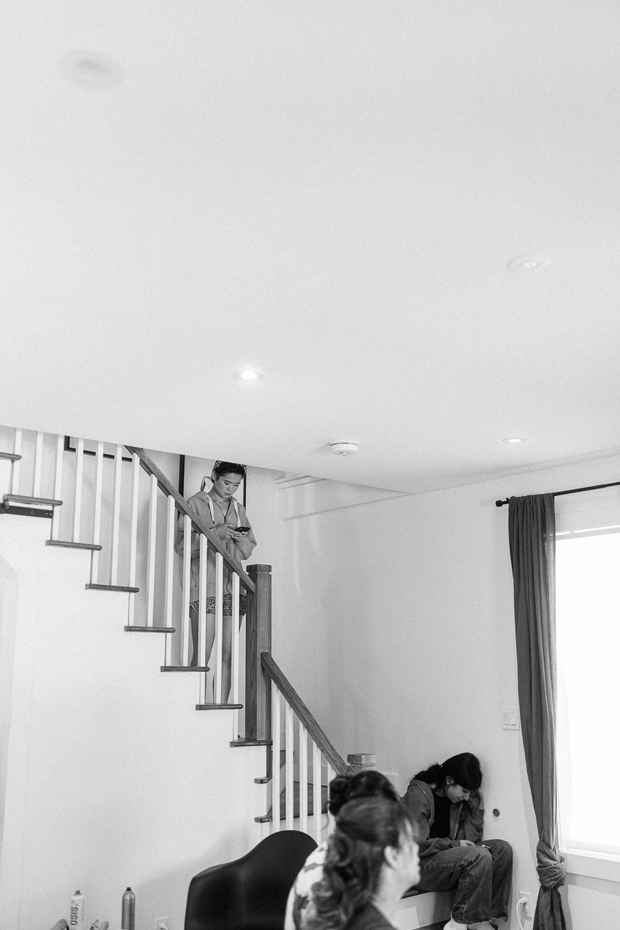 Black-and-white scene of people waiting on a staircase and by a window during wedding preparations.