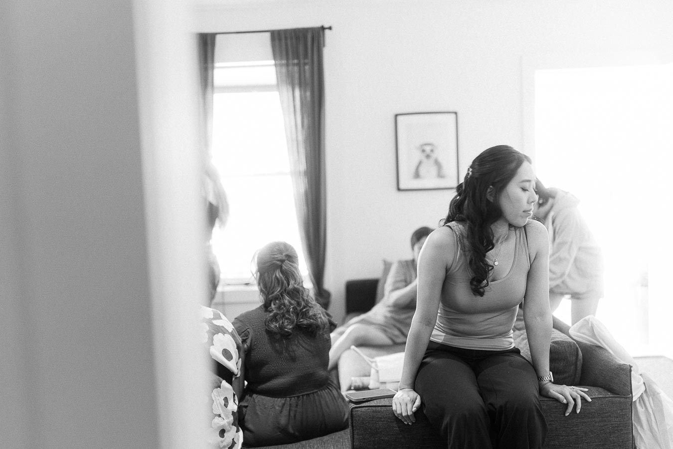 Black-and-white scene of a woman sitting on a sofa while others relax in a bright getting-ready room.