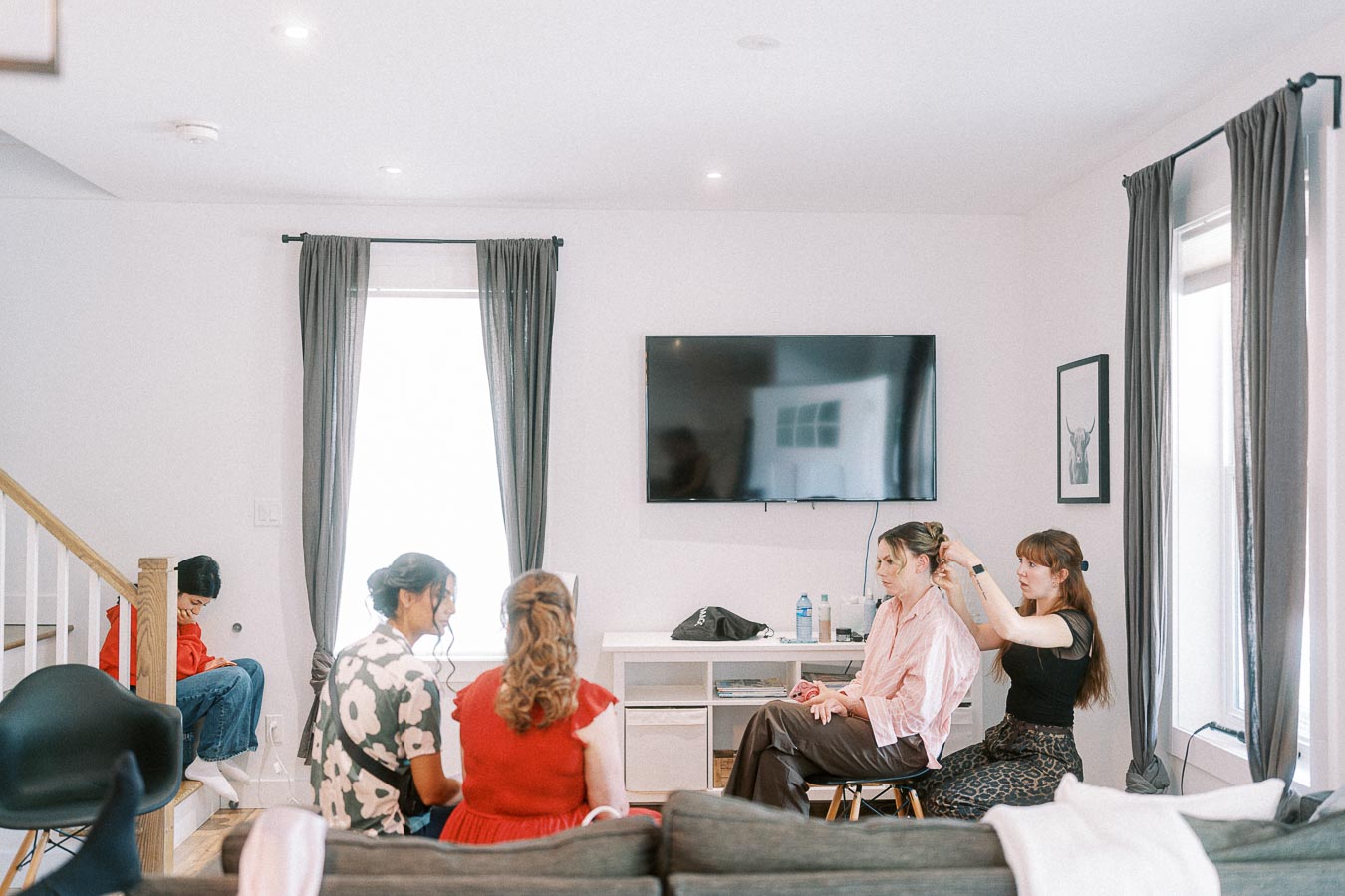 Bride sits in a bright room while a stylist adjusts her hair and others relax nearby.