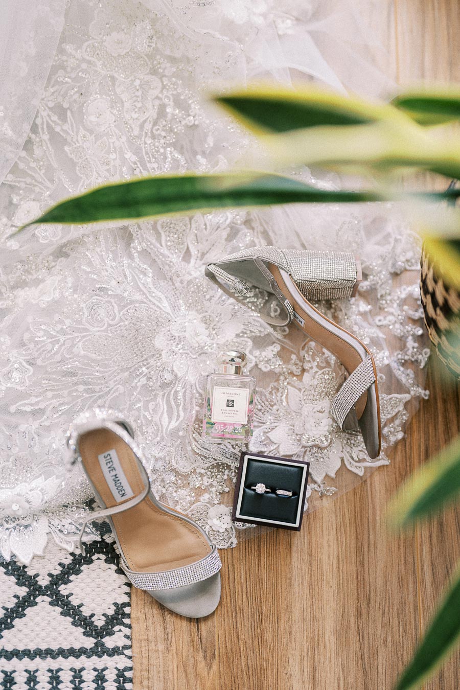 Rhinestone heels, perfume, and an engagement ring arranged on a lace wedding dress with greenery in front.