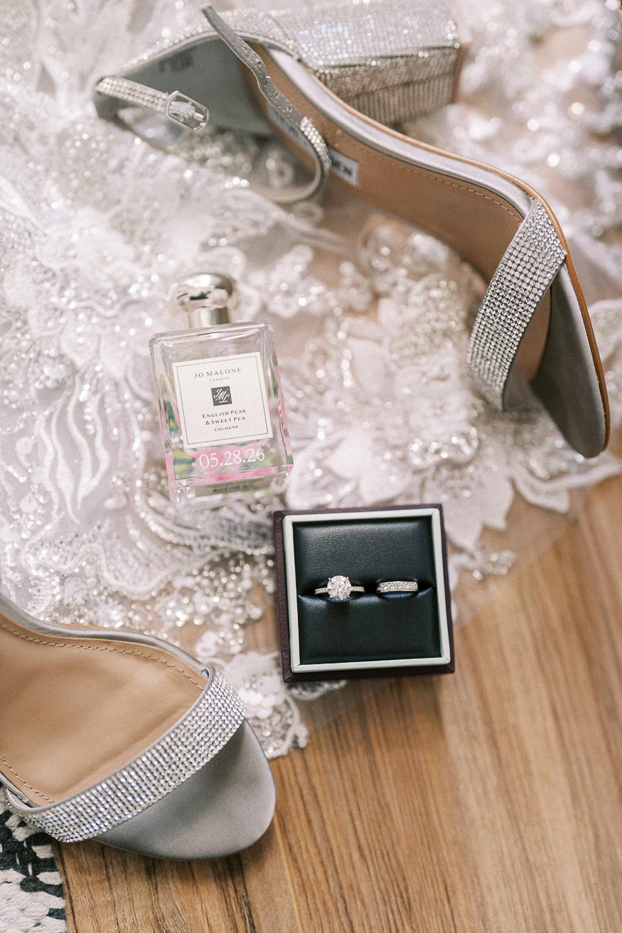 Silver bridal heels, Jo Malone perfume, and wedding rings arranged on a lace gown with beading.