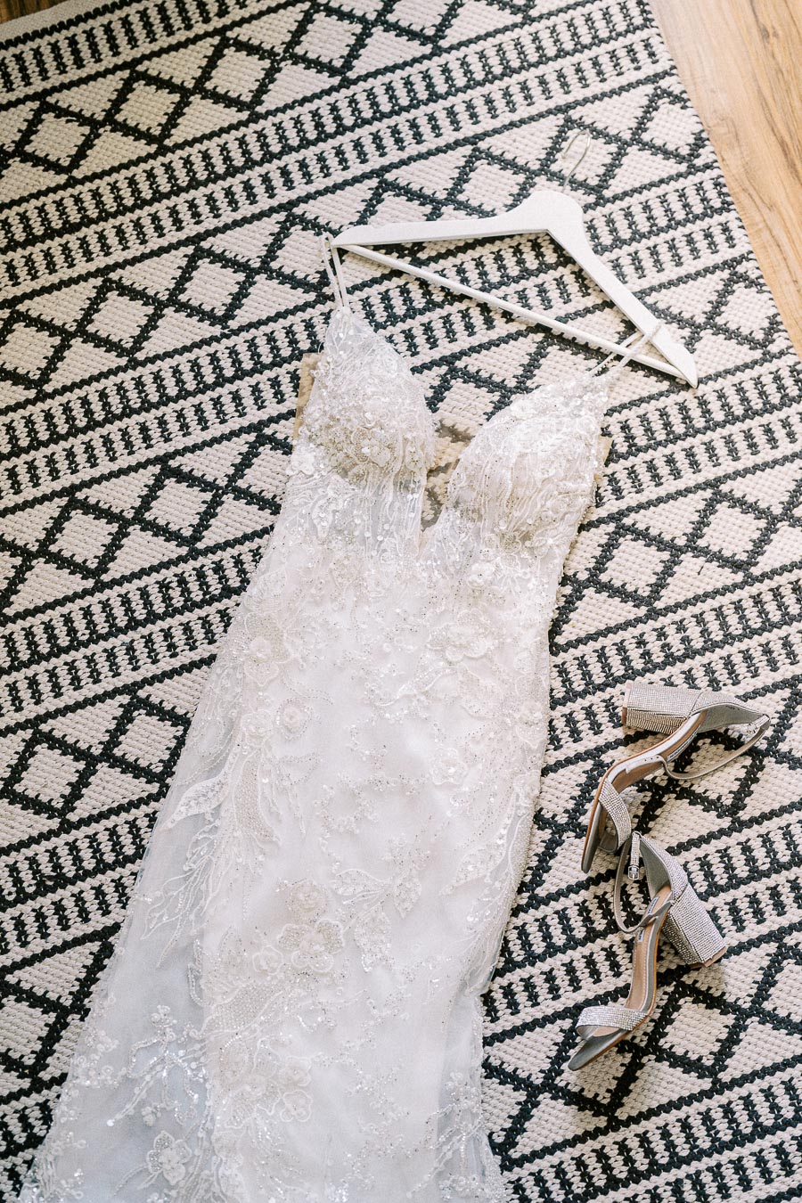 Beaded lace wedding dress on a white hanger with silver heels on a black and white patterned rug.