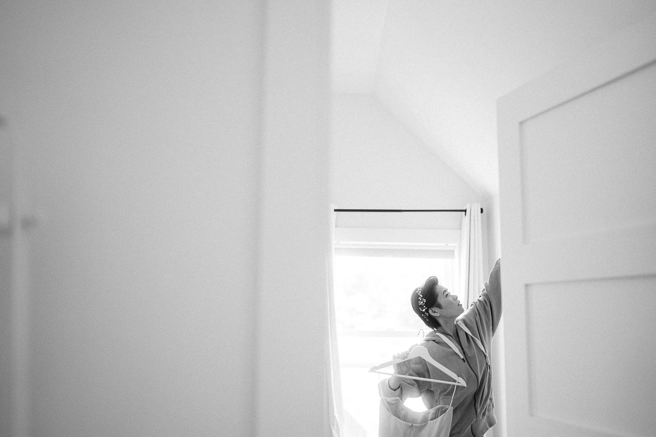 Bride in a robe reaches upward beside a window while holding a dress hanger in a bright room.
