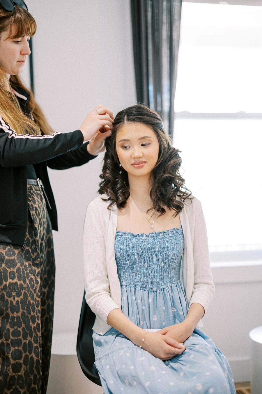 Woman seated by a bright window while a stylist pins curls into her hair during wedding morning preparations.