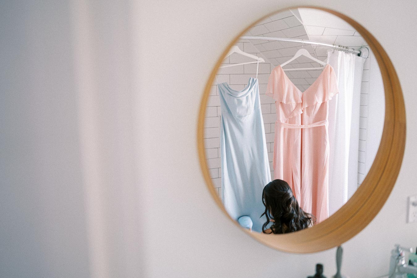 Round mirror reflecting pastel bridesmaid dresses hanging above a seated woman in a bright getting-ready room.