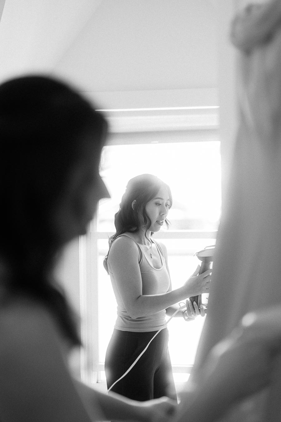 Woman uses a garment steamer on a hanging wedding dress beside a bright window during preparations.