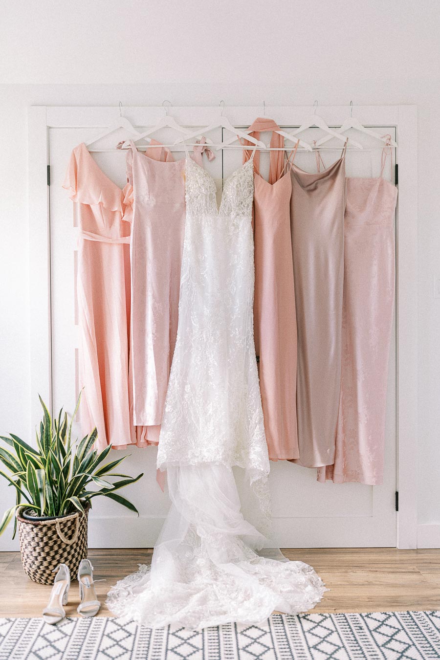 White lace bridal gown hanging among blush bridesmaid dresses, with heels and a potted plant on the floor.