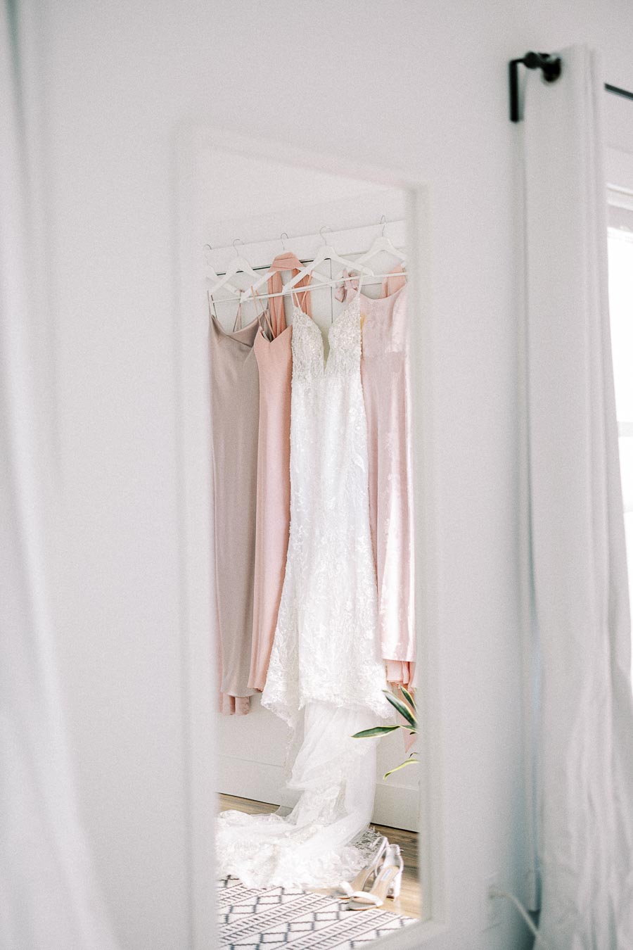 Lace wedding dress and pastel bridesmaid dresses hanging by a mirror with bridal shoes below