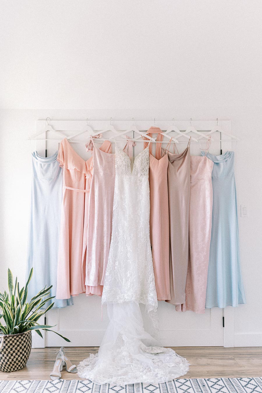 Lace wedding dress with pastel blue, pink, and taupe bridesmaid dresses hanging on a white wall rack.