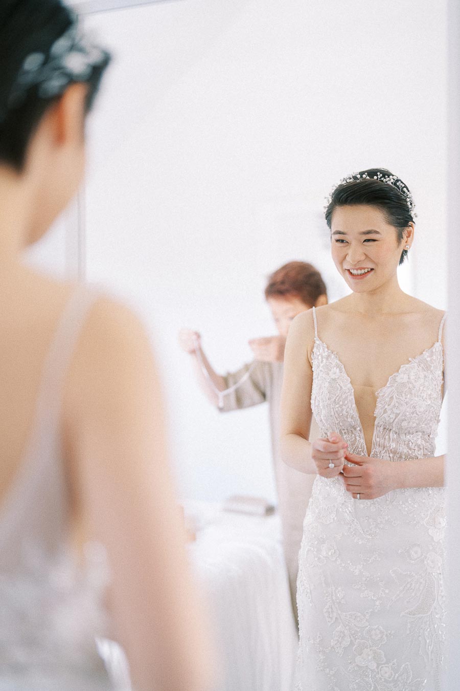 Bride smiling in a mirror while wearing a beaded lace wedding dress and jeweled headband during getting ready