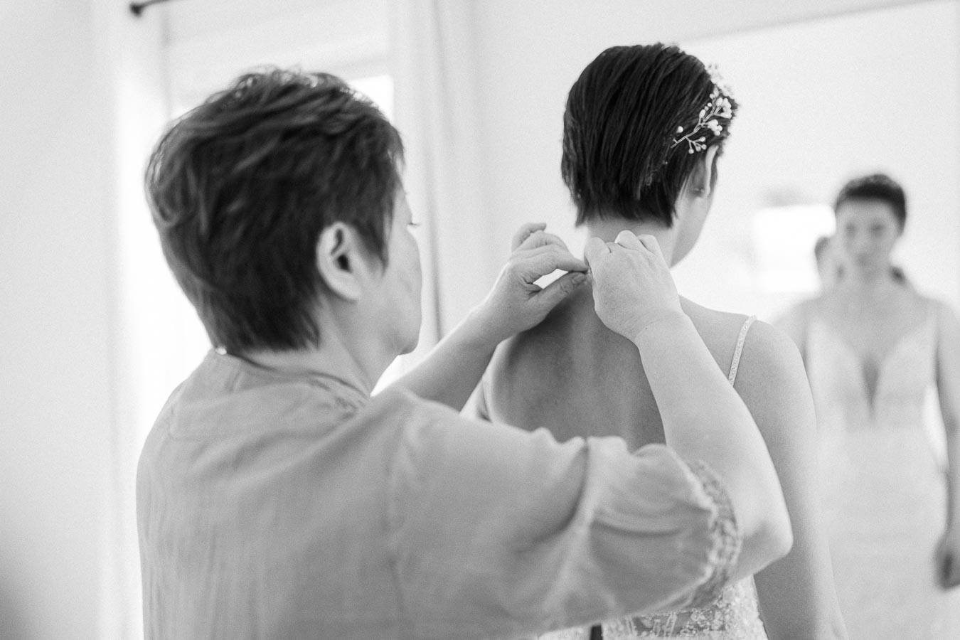 Woman fastens the back of Miffy's wedding dress as another bridesmaid watches in a softly lit room.