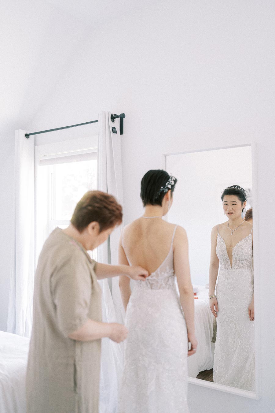 Bride in a lace wedding dress looks in a mirror while another person fastens the back in a bright room.