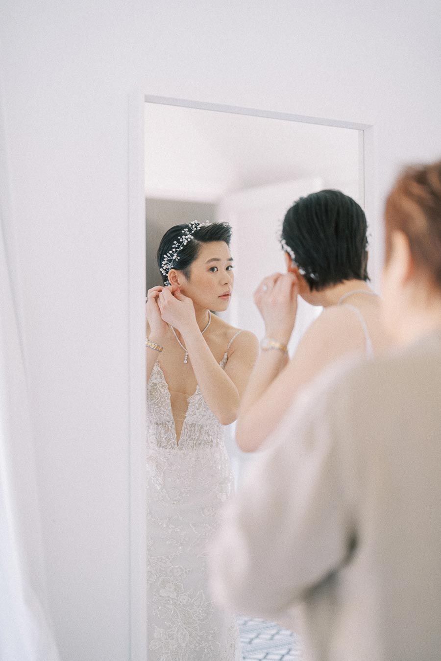 Bride in a beaded gown adjusts her earring while looking into a mirror during wedding preparations.