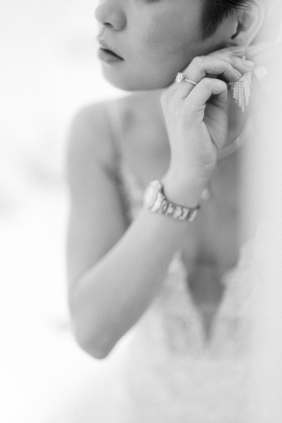 Bride fastening a dangling earring while wearing an engagement ring and watch in a soft black-and-white close-up.