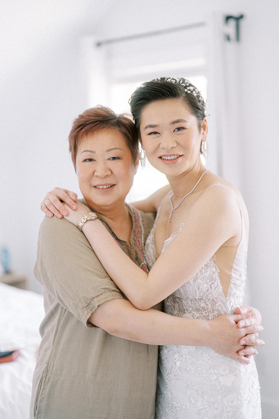 Bride Miffy in a lace wedding dress hugs an older woman in a bright getting-ready room.