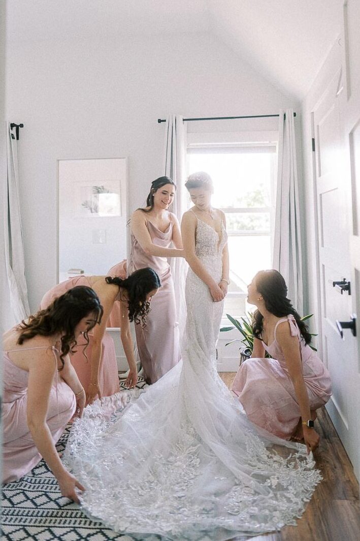 Bride in a lace gown stands as bridesmaids in pink dresses arrange her train in a bright room.
