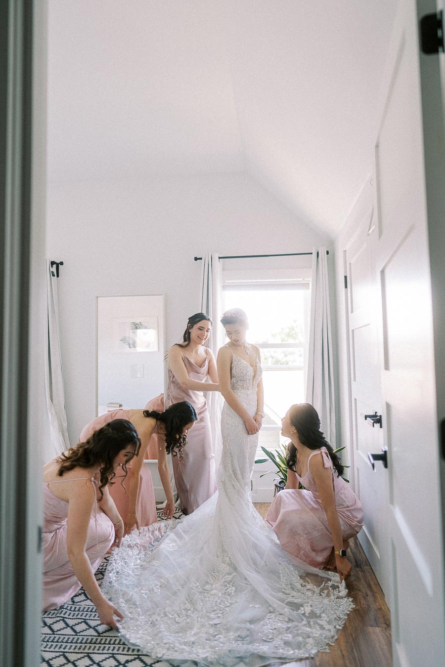 Bride in a lace gown stands as bridesmaids in pink dresses arrange her train in a bright room.