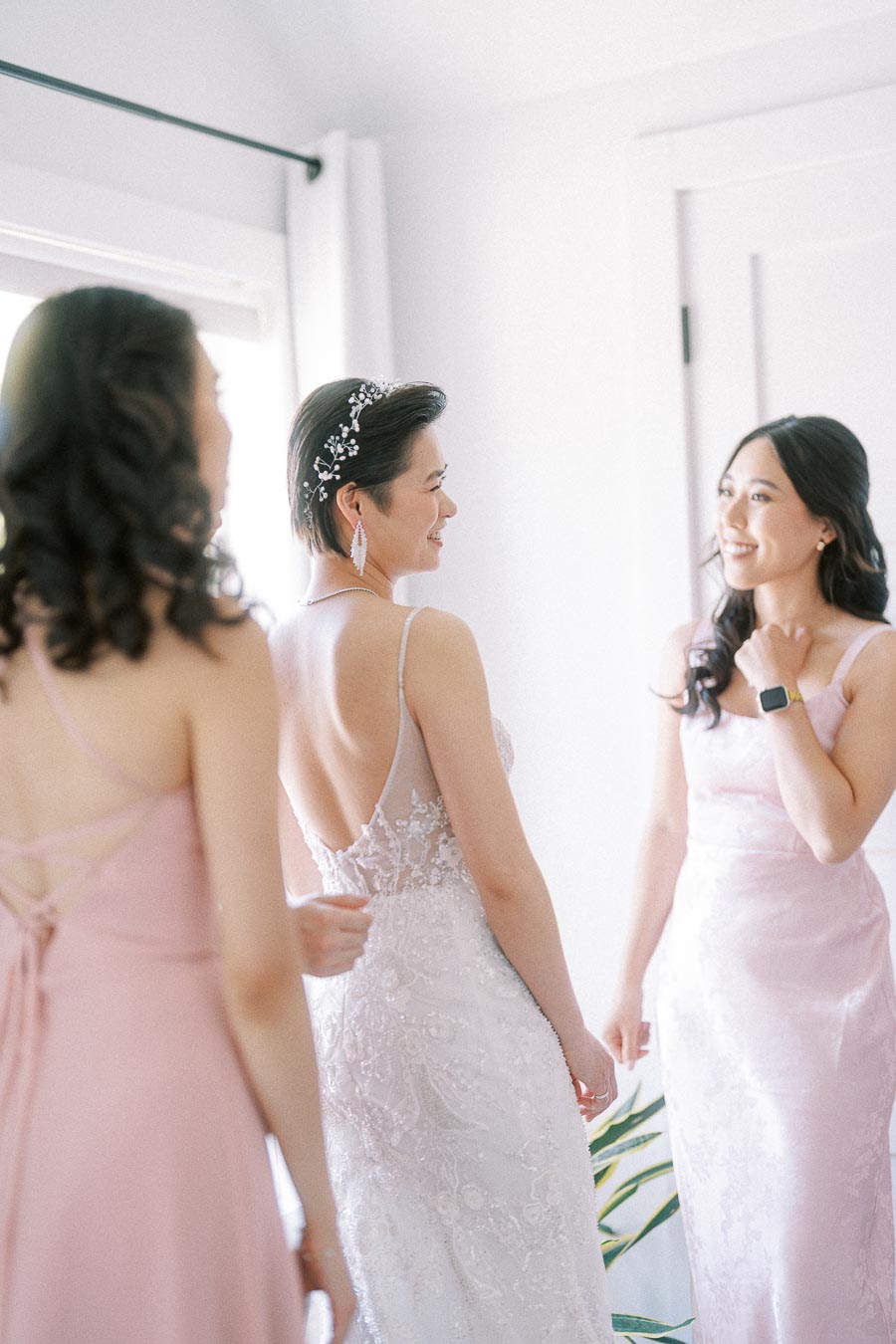 Bride in a lace gown smiles with two bridesmaids in pink dresses during wedding morning preparations.