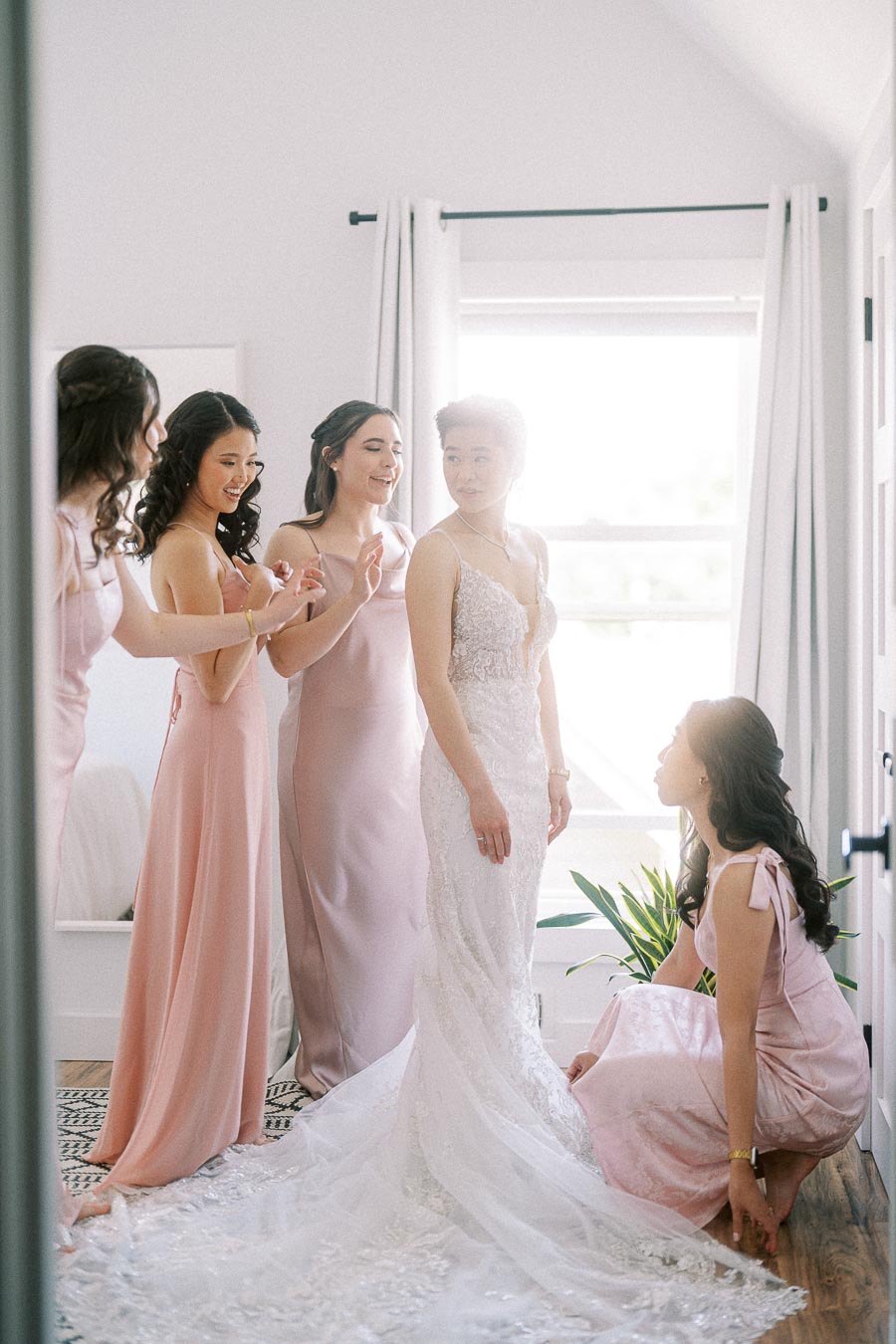 Miffy stands in a lace wedding gown as bridesmaids in pink dresses adjust her train in a bright room.
