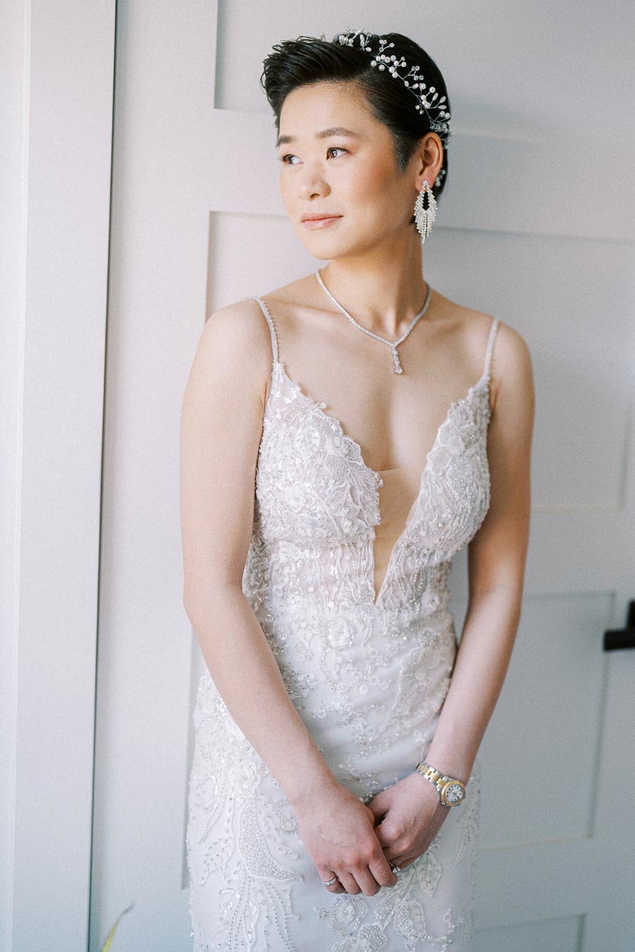 Bride in a beaded lace gown with jeweled headpiece stands by a window looking to the side.