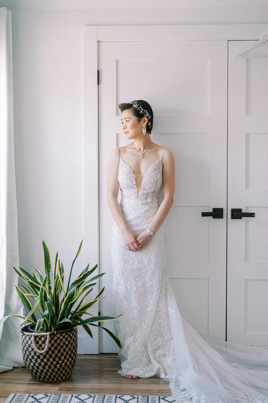 Miffy stands by a window in a lace wedding gown with a long train, floral hairpiece, and potted plant nearby.