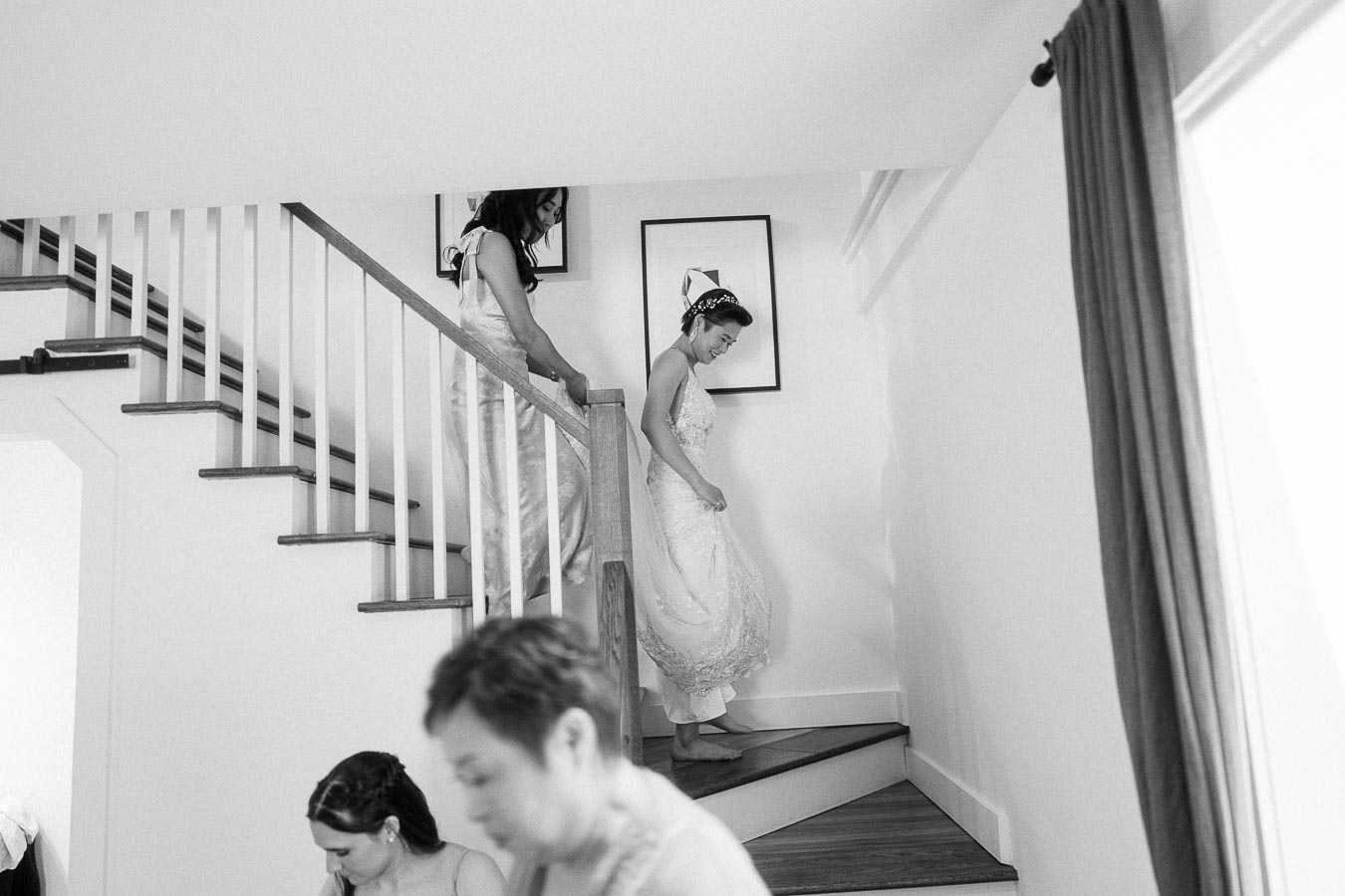 Bride in a lace dress descends an indoor staircase with a bridesmaid nearby during wedding preparations.