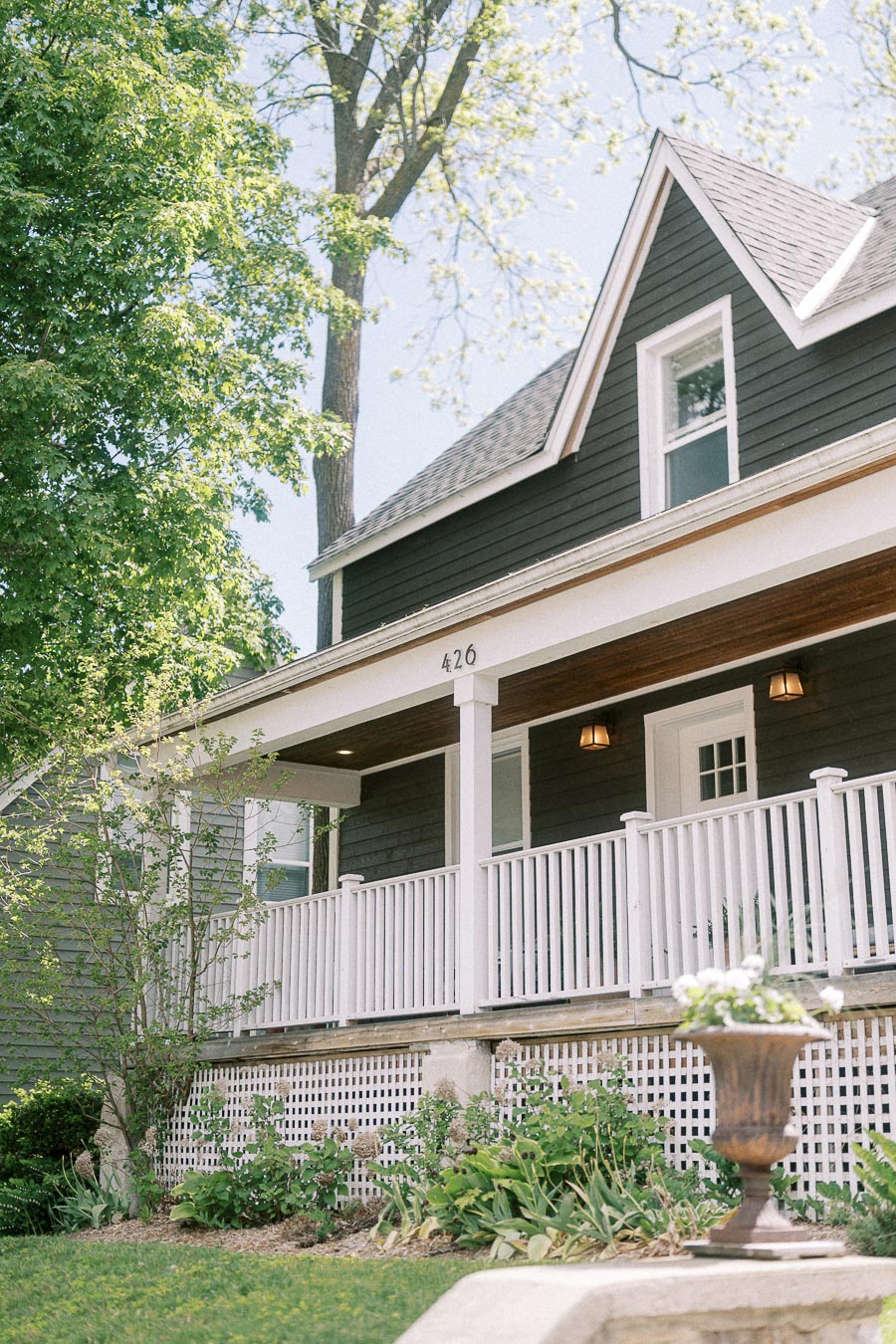 Black house with white porch railings, number 426, garden greenery, and trees in bright daylight.