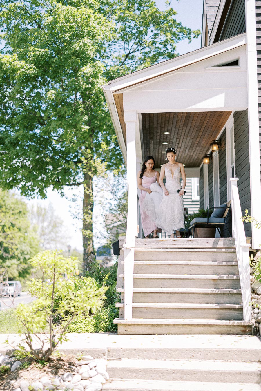 Bride in a lace wedding dress walks down porch steps with a bridesmaid in a pale pink dress beside her.