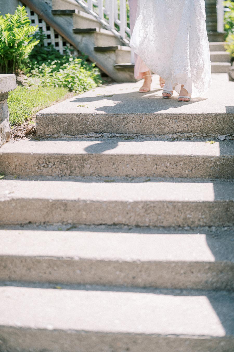 Bride in a lace wedding dress stepping down sunlit stone stairs with garden greenery nearby.
