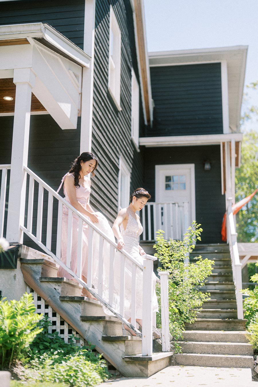 Bride in a white gown and attendant in pale pink walking down outdoor stairs beside a dark building
