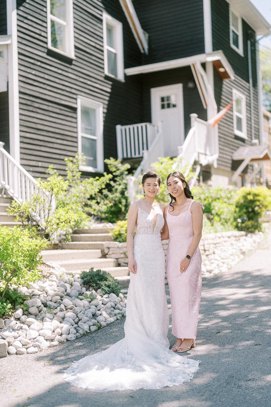 Bride in a lace wedding gown poses with a woman in a pale pink dress outside a black-and-white building.