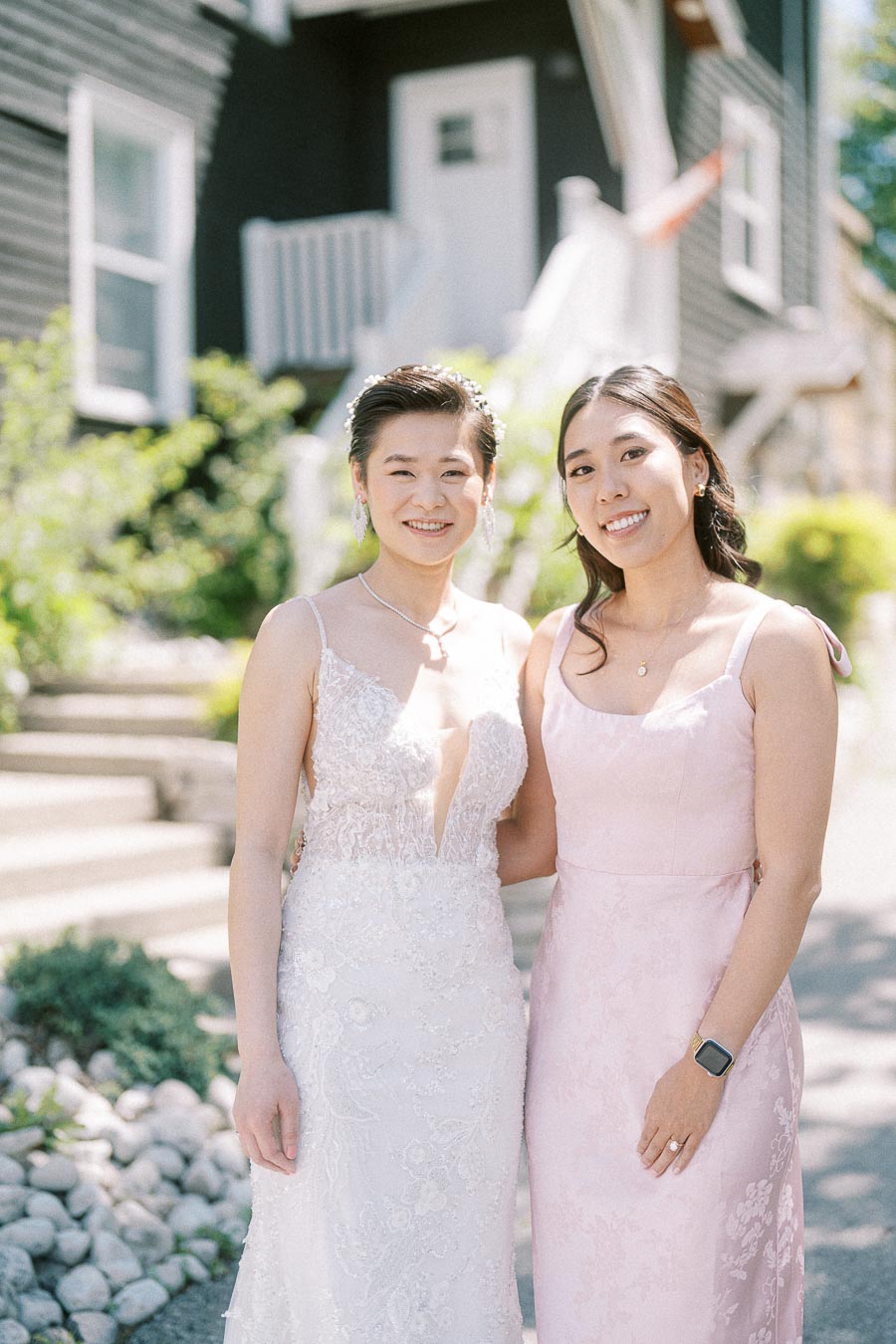 Bride in a lace gown stands with a smiling woman in a pale pink dress outside Ancaster Mill.
