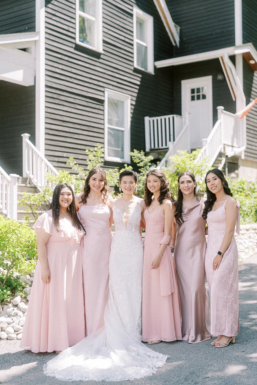 Bride in a lace gown stands with five bridesmaids in blush dresses outside a dark clapboard building.