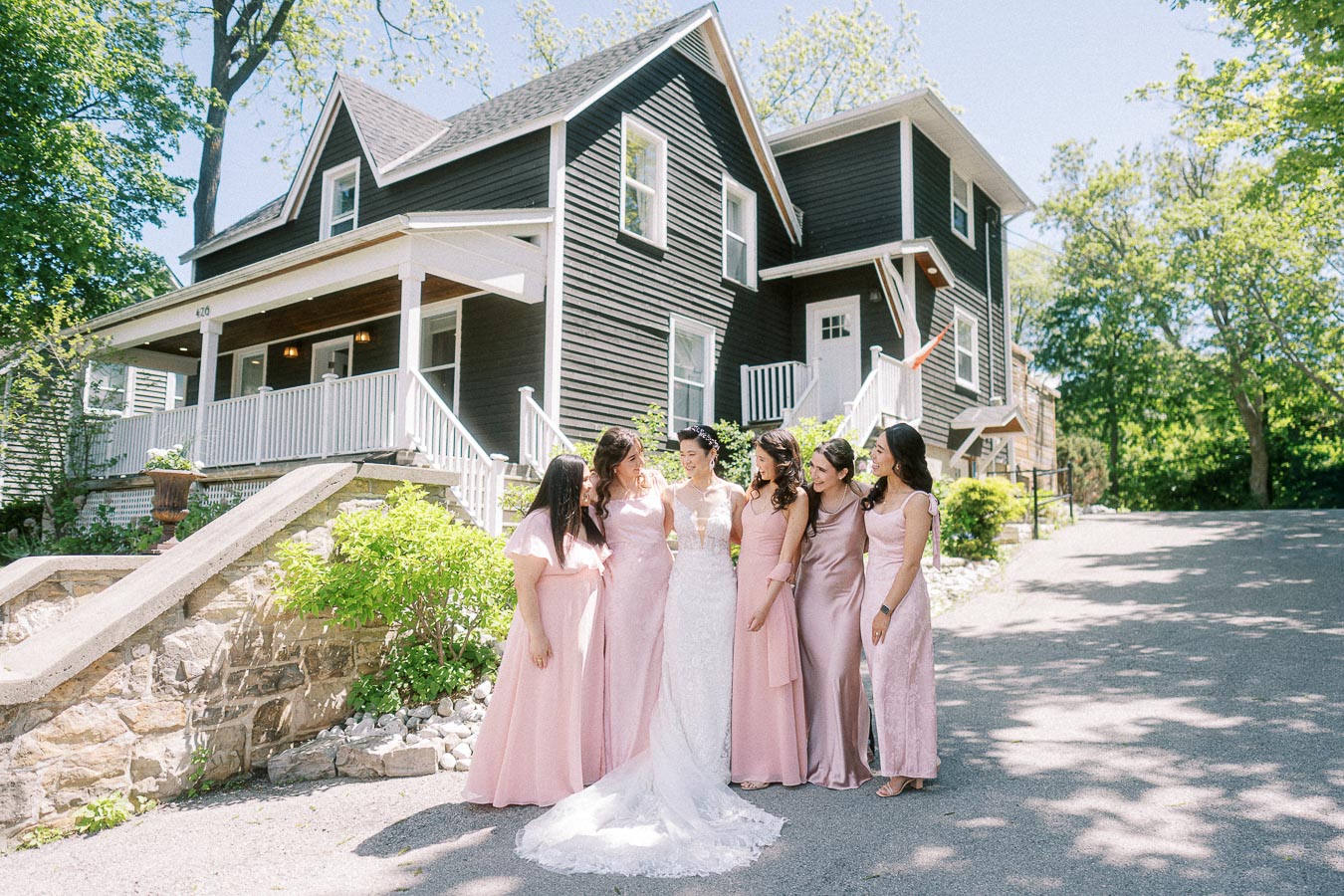 Bride in a lace gown stands with five bridesmaids in pink dresses outside a black house at Ancaster Mill.