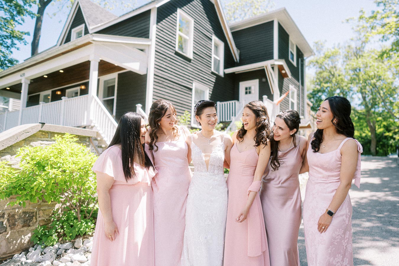 Bride in a white gown laughing with five bridesmaids in pink dresses outside a dark house at Ancaster Mill
