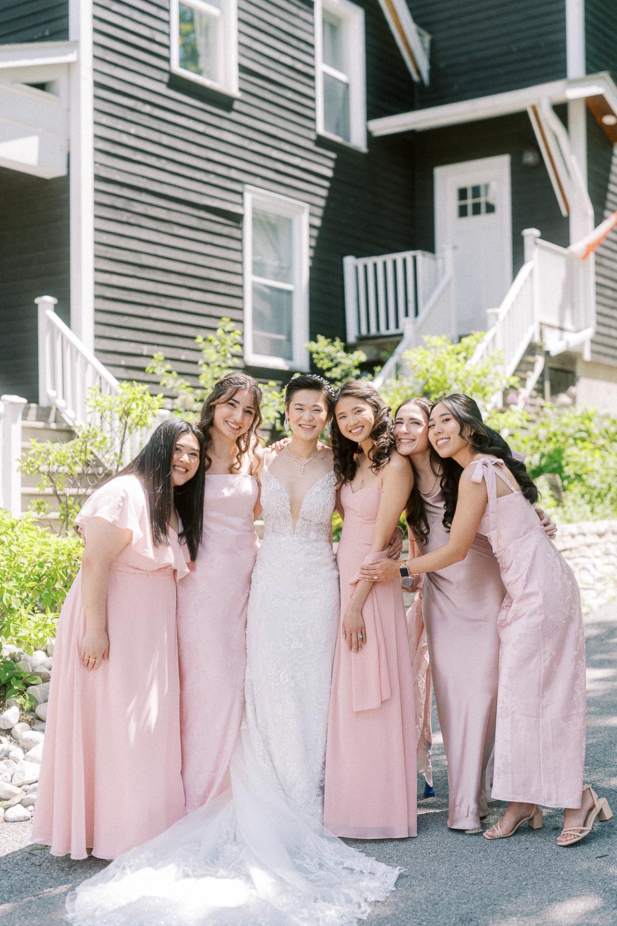 Miffy in a white lace wedding gown poses with five bridesmaids in pale pink dresses outside a dark wood venue.