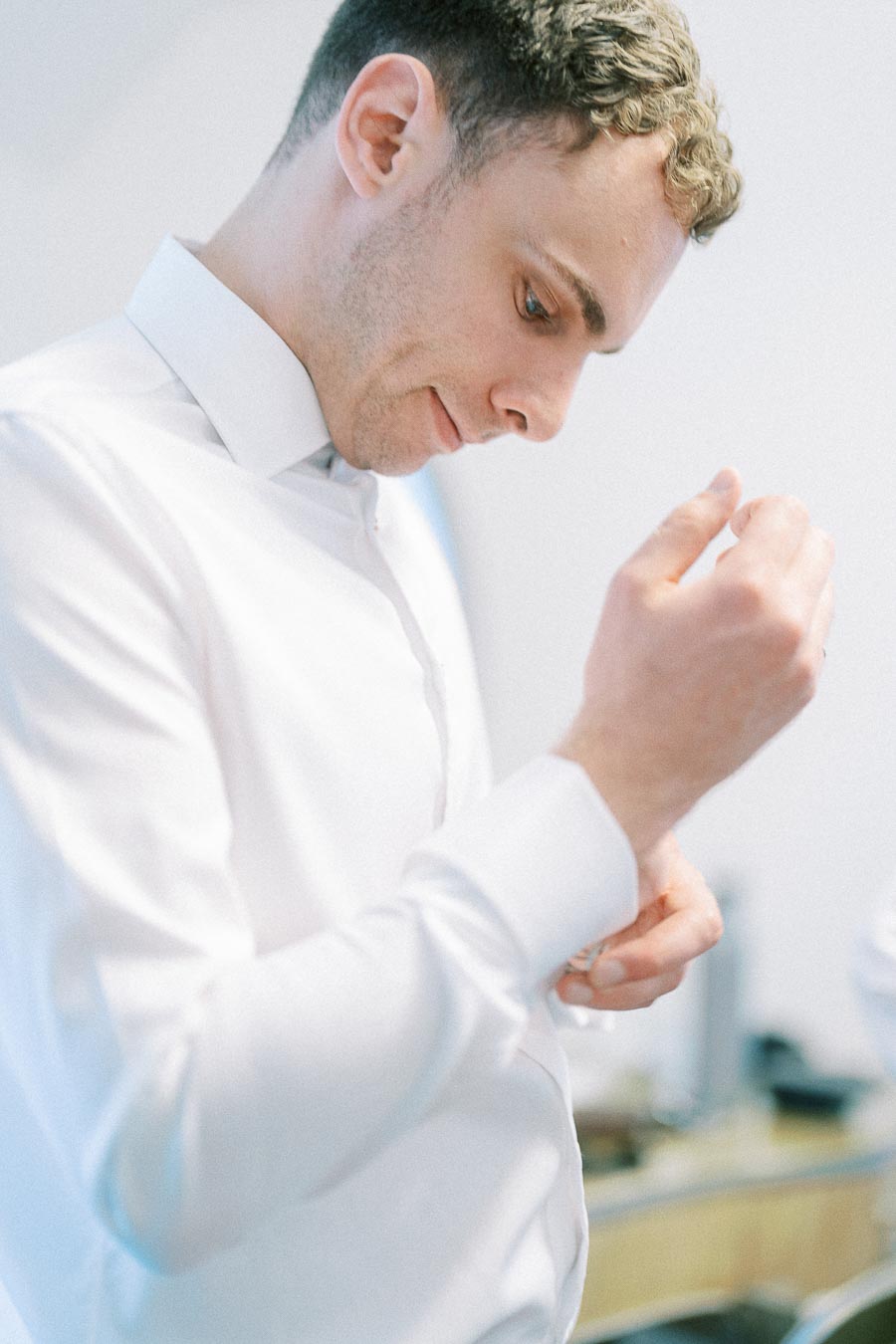 Groom in a white dress shirt fastening his cuff while getting ready in soft window light.