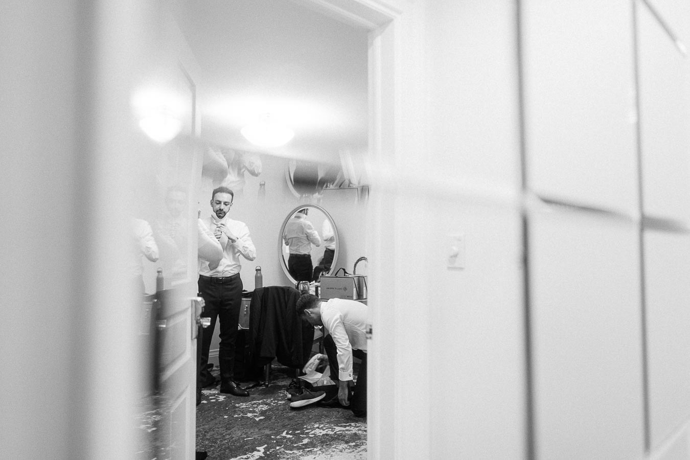 Groom in a white shirt prepares with another man adjusting shoes in a small getting-ready room.