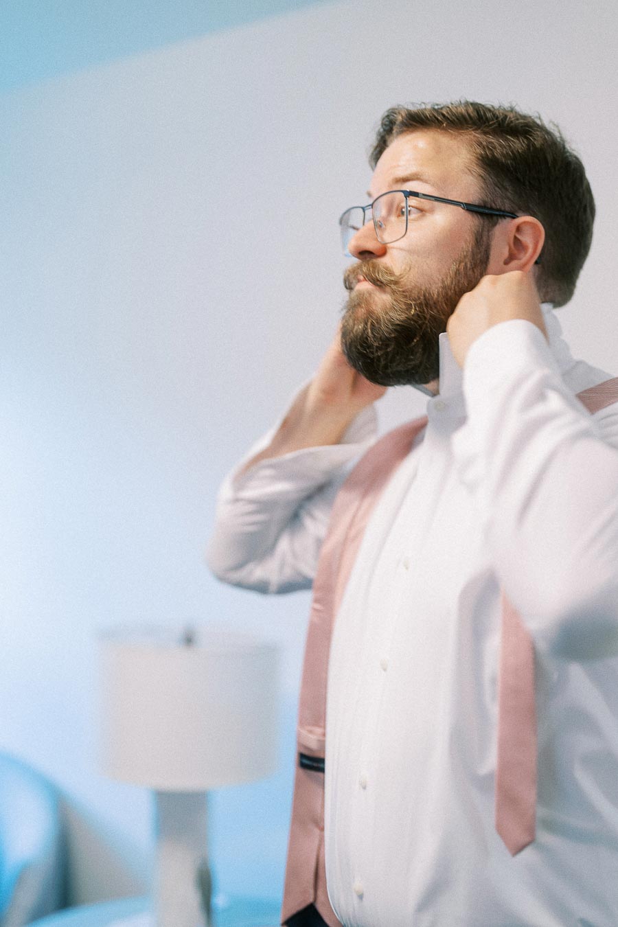 Man with glasses adjusts his collar while wearing a white shirt and pale pink suspenders indoors.