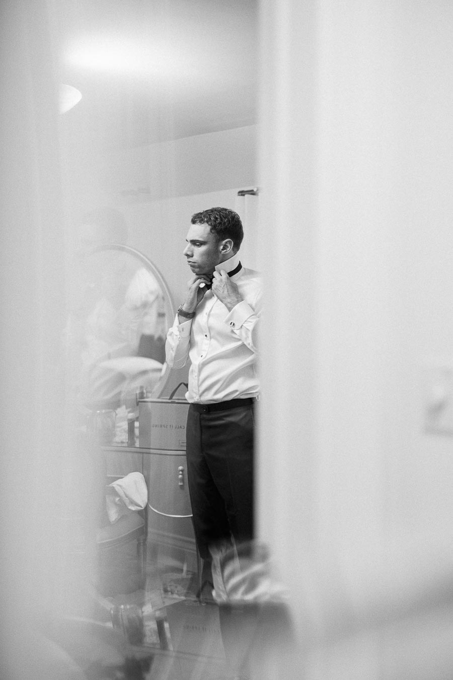 Groom adjusting his black bow tie in a white dress shirt while getting ready indoors.