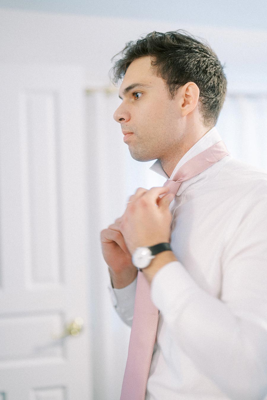 Rawle adjusts a pale pink tie while wearing a white dress shirt and watch in a bright room.