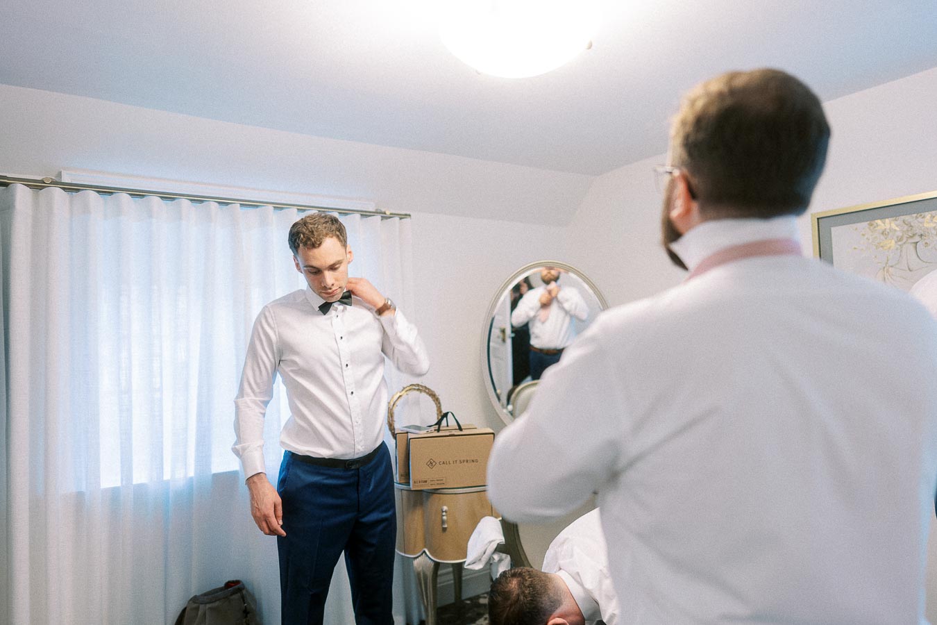 Groom in a white shirt adjusts his collar while another man gets ready in a bright room.