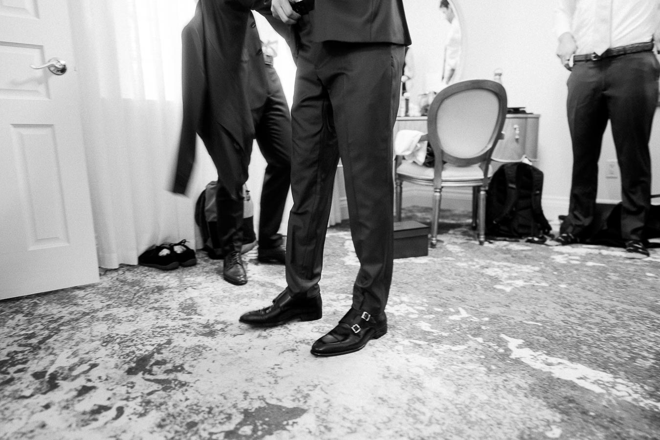 Groomsmen in dress shoes and trousers getting ready in a room with chairs, bags, and soft window light.