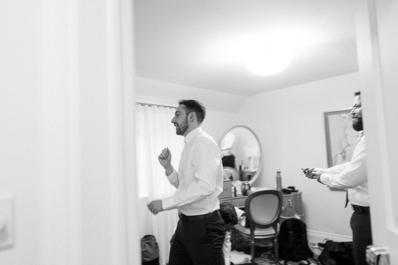 Two men in dress shirts getting ready in a bright room, one adjusting his cuff near a vanity mirror.