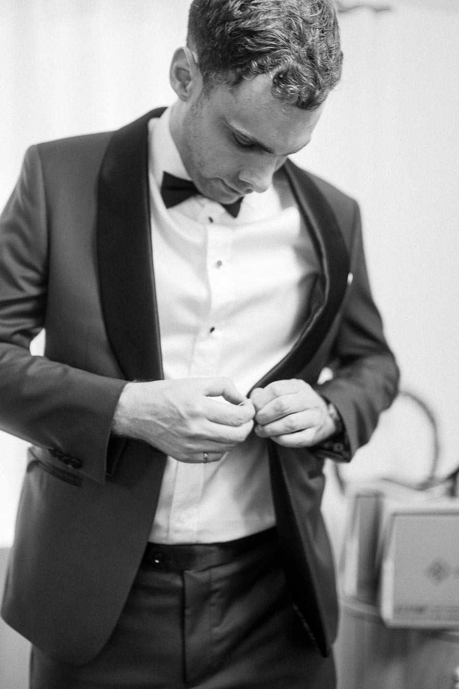 Groom in a black tuxedo fastening his jacket while looking down during wedding preparations.