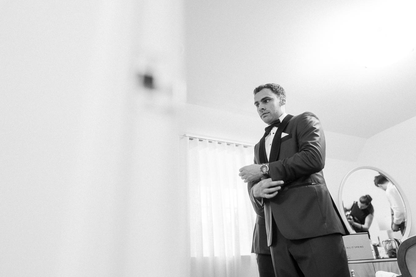 Groom in a tuxedo adjusts his cuff in a bright room, with people reflected in a round mirror behind him.